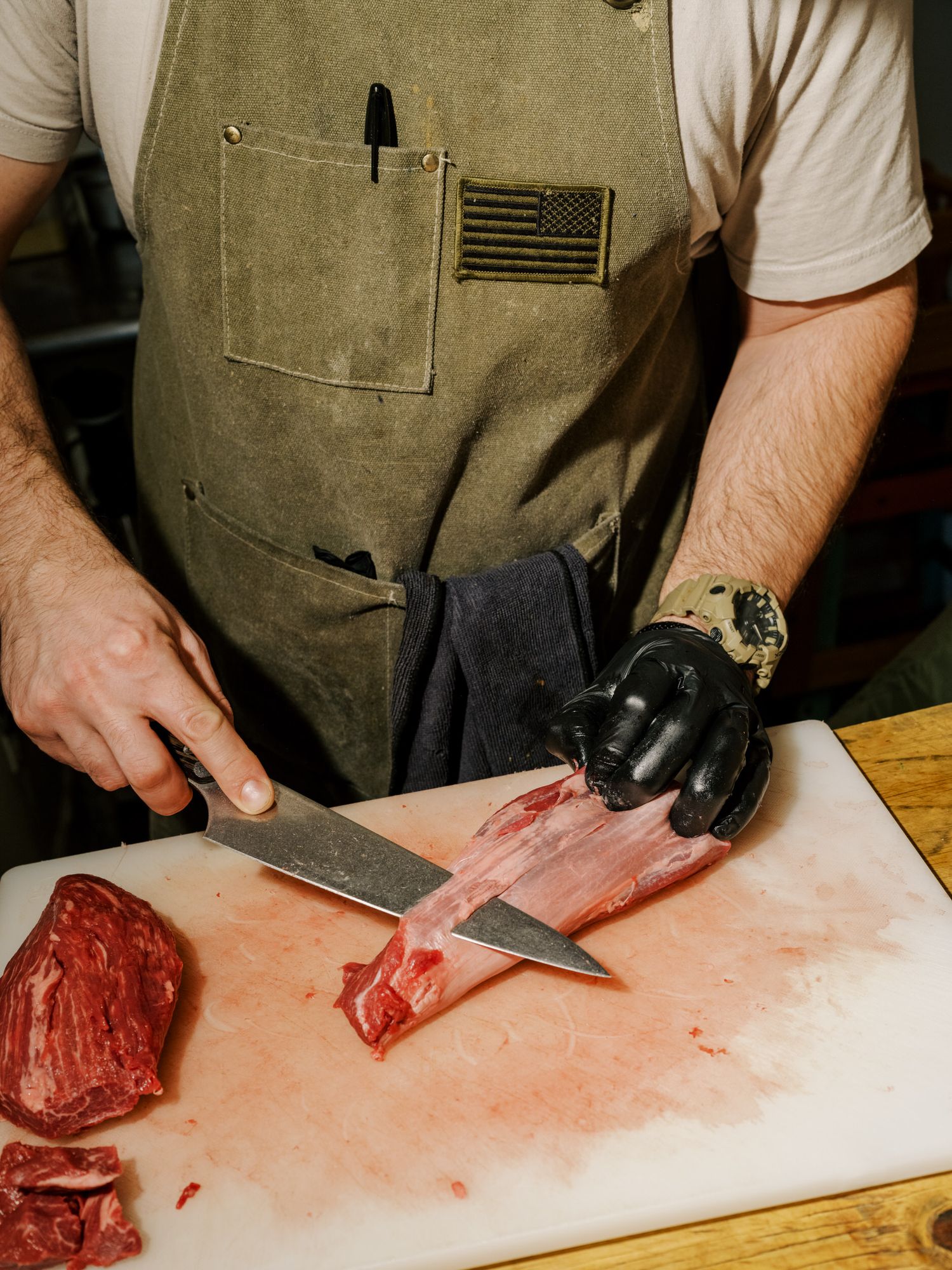 Professional butcher in olive apron cuts fresh raw meat on white cutting board while wearing black gloves.