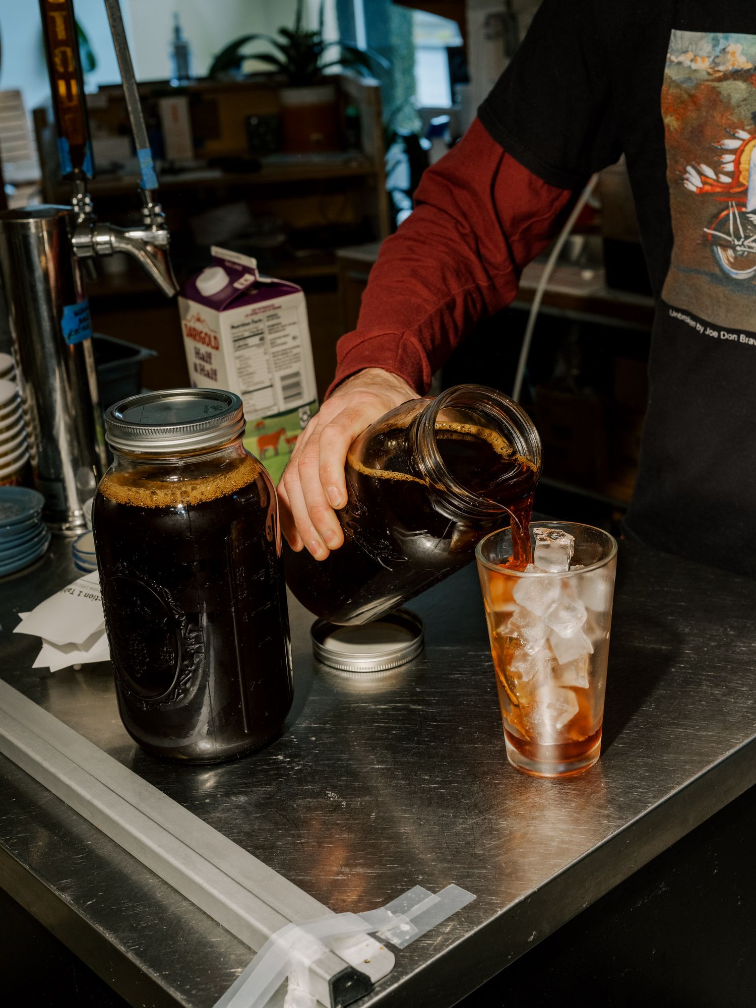 Person pouring cold brew coffee from a large mason jar into a glass filled with ice at a coffee shop counter.