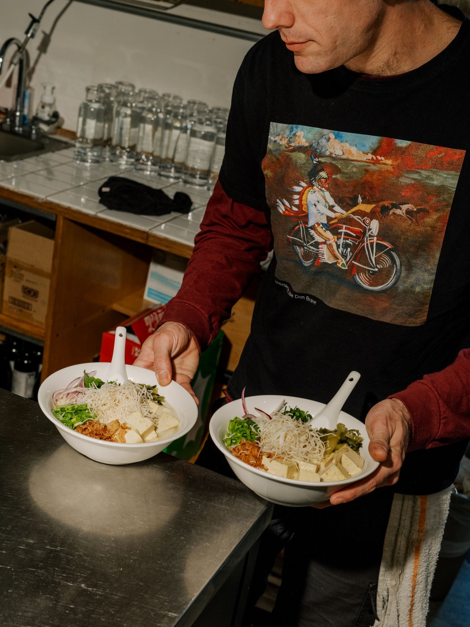 Two bowls of Asian noodle soup with garnishes being served on dark countertop by someone wearing a graphic t-shirt.