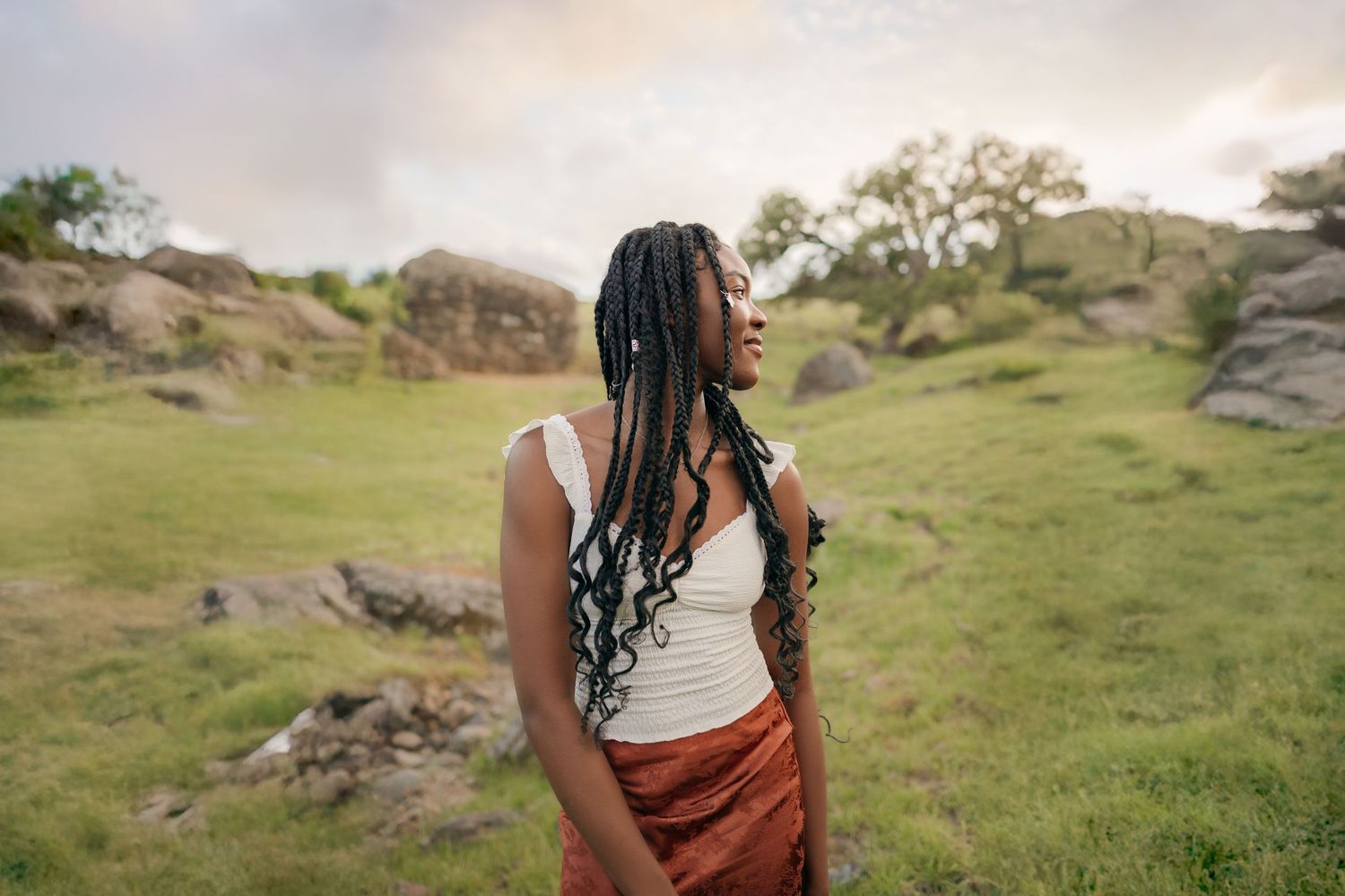 Senior portrait of black female student with long braids, white tank, and orange skirt gazing off camera with rolling hills/trees/rocks