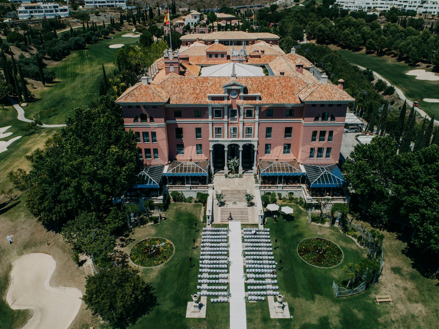 Aerial view of a grand pink Mediterranean-style mansion with formal gardens and outdoor wedding ceremony setup.