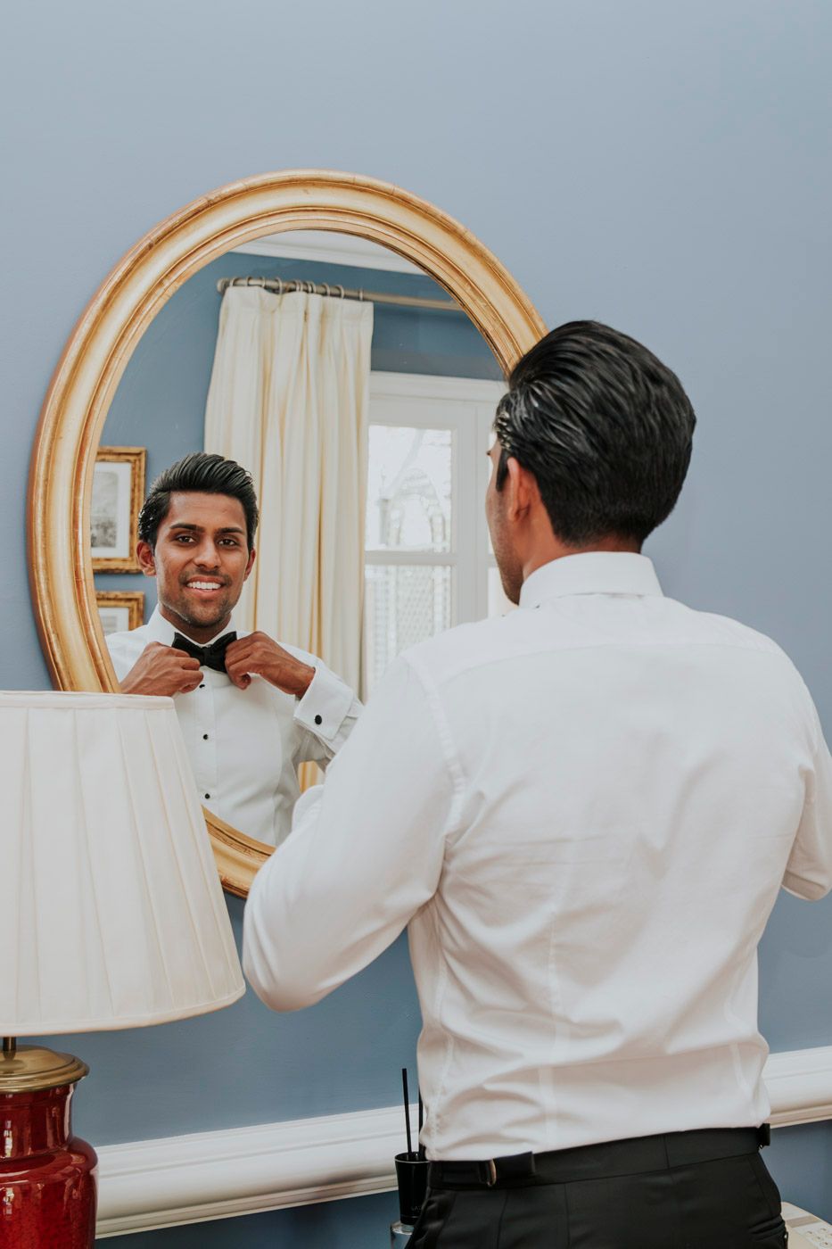 A reflection in an oval gold-framed mirror shows someone adjusting a black bowtie while wearing formal attire.