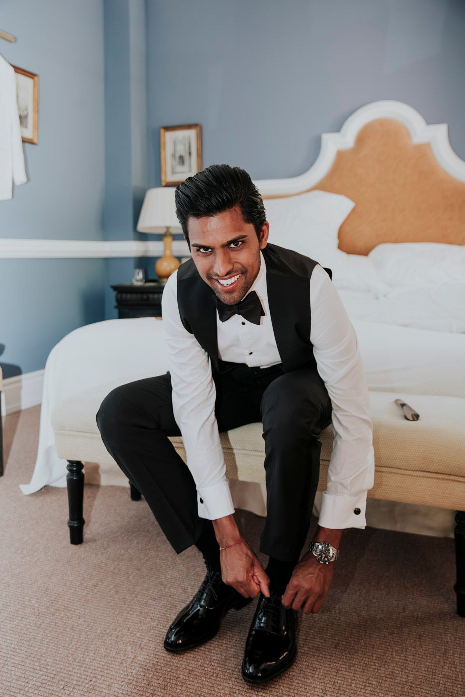 Someone in a classic black tuxedo getting ready, putting on formal shoes while sitting on a bed in a blue-walled room.