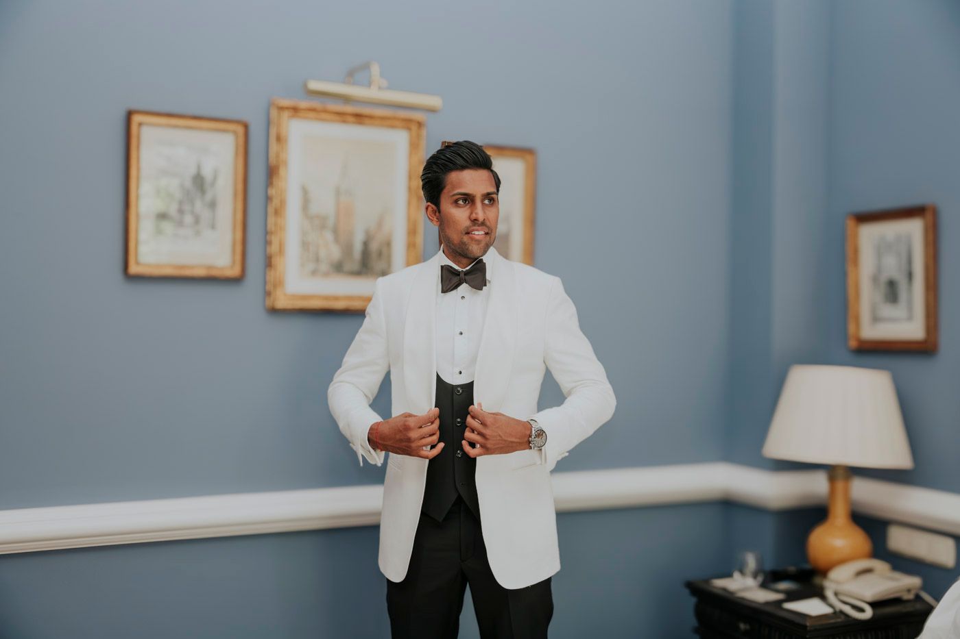 A person wearing a white dinner jacket with black bow tie stands in an elegant room with framed artwork and a table lamp.
