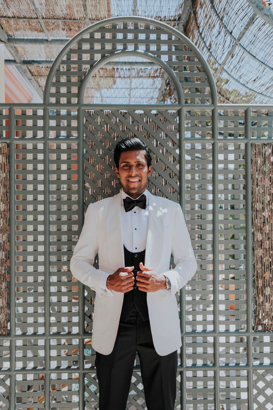 A person wearing a white dinner jacket and black bow tie stands in front of a latticed garden arbor.