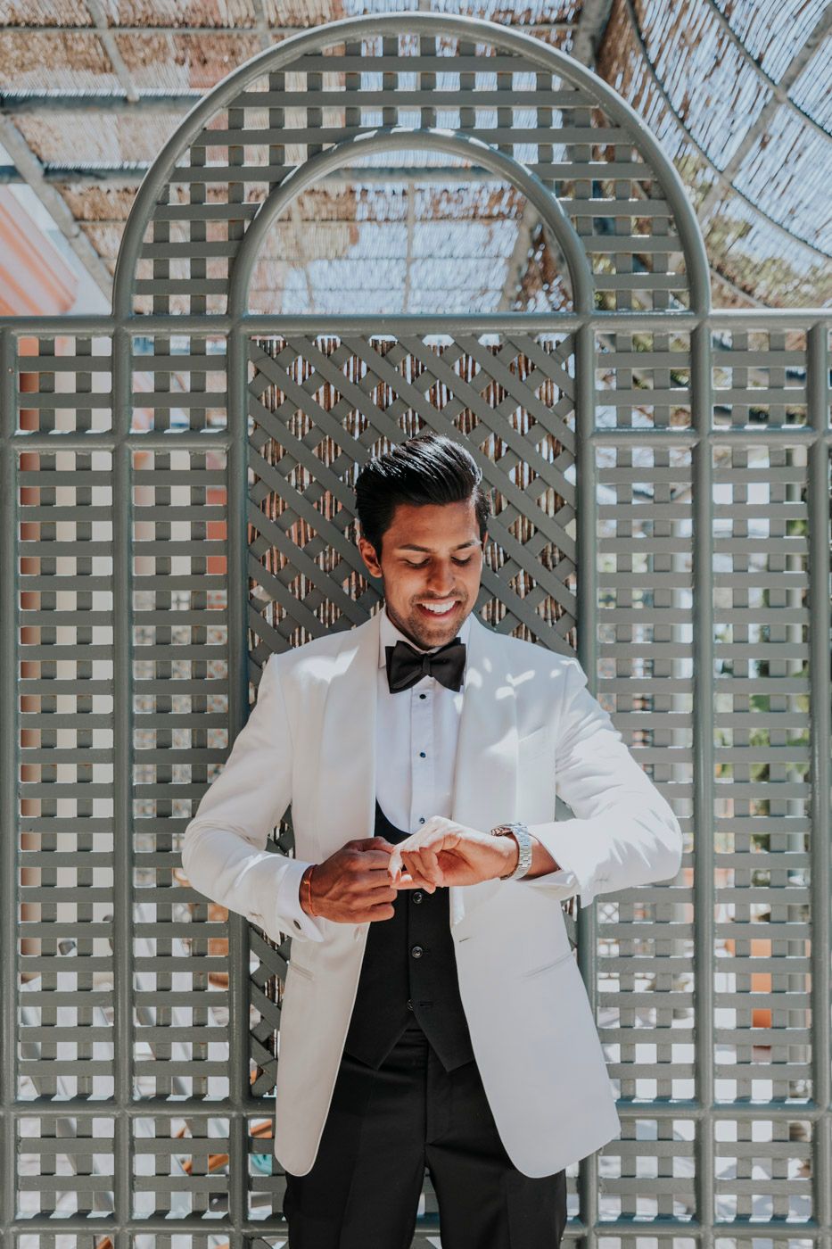 A formal dressed person checks their watch while standing against a gray latticed backdrop.
