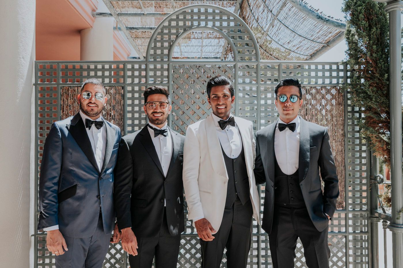 Four people in formal attire stand together under a garden arbor for a wedding photo.