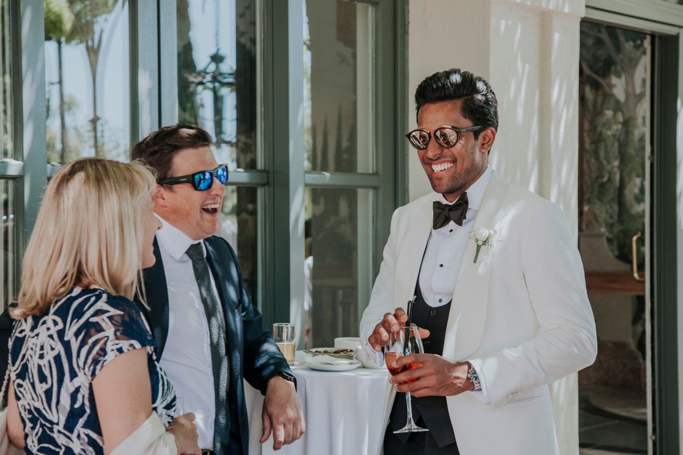 Guests in formal attire socialize at an elegant outdoor cocktail reception with white tables and palm trees.