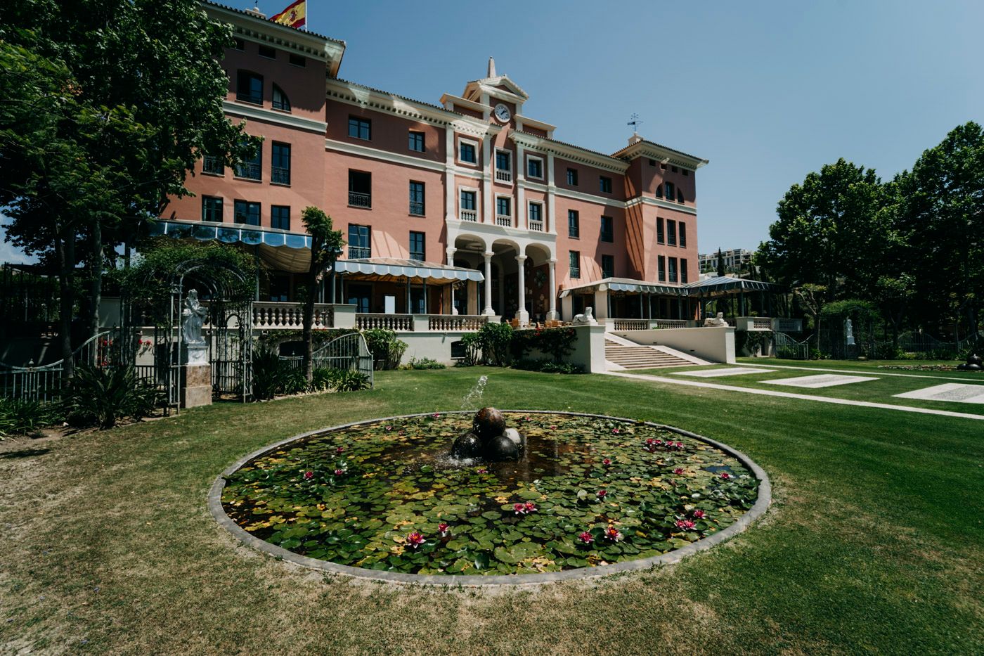 Grand historic mansion with pink facade and manicured gardens featuring a circular floral display on a sunny day.
