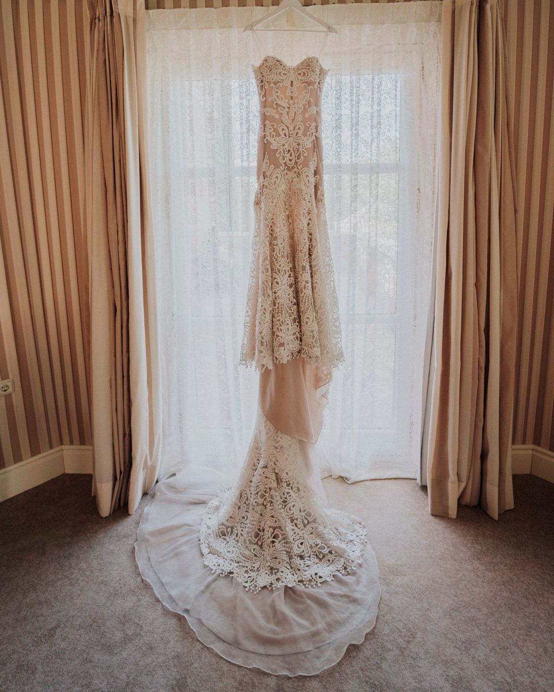 Elegant lace wedding dress with long train hanging in front of sheer curtains and brown drapes.