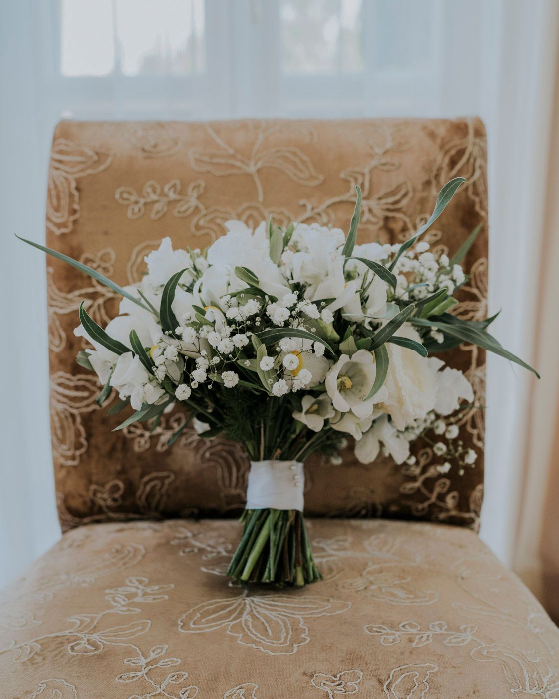 White floral wedding bouquet with roses and baby's breath on a beige textured chair.