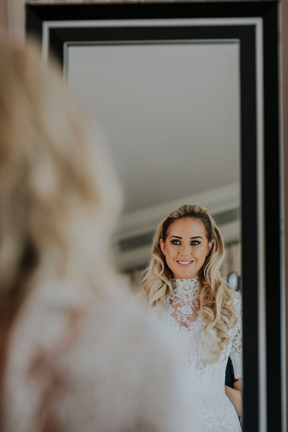 A person in a wedding dress with long wavy blonde hair looks at their reflection in an ornate black framed mirror.