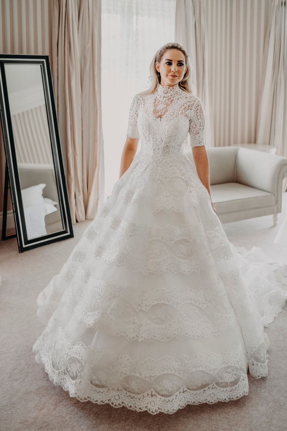An intricate white lace wedding gown with high neck, half sleeves and full skirt shown in a bridal fitting room.