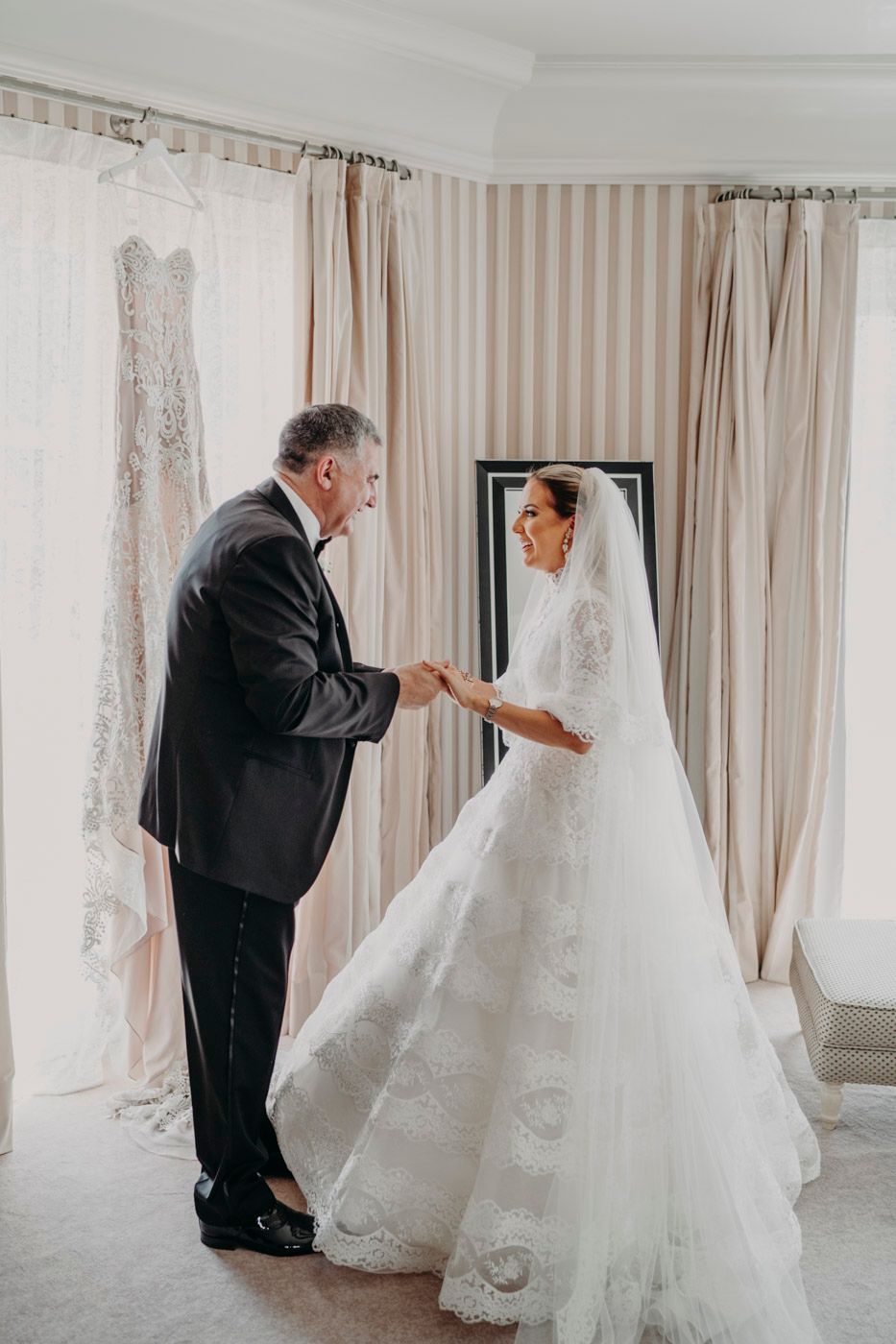 A tender moment during wedding preparations with a flowing white lace gown in the background.
