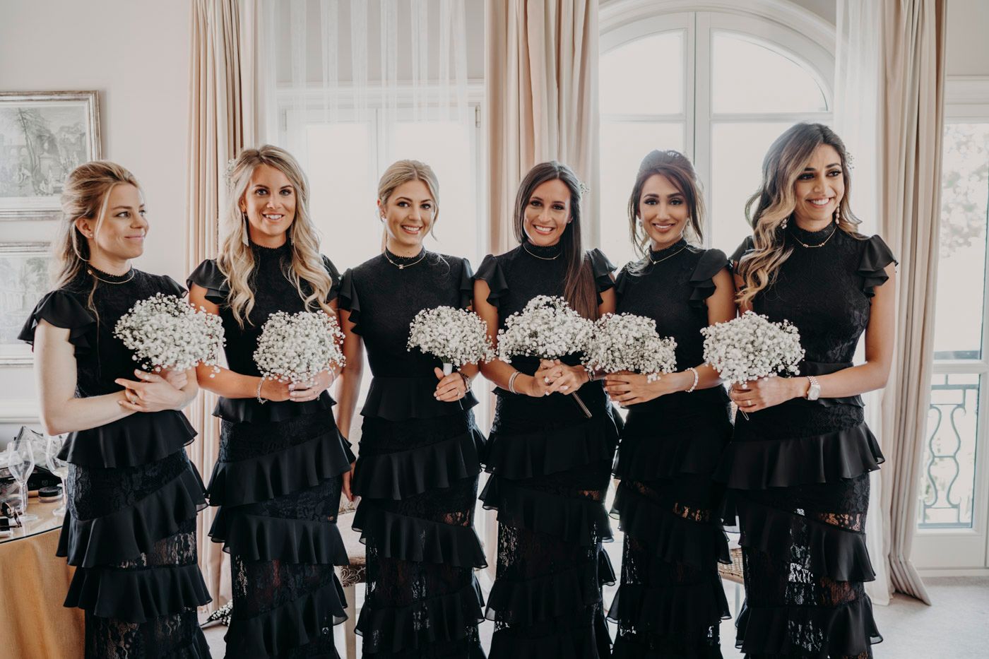 A group of bridesmaids wearing matching black lace gowns hold white floral bouquets in an elegant room.