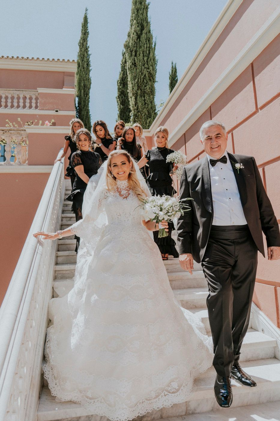 Wedding party descends elegant pink marble staircase at outdoor venue with cypress trees in background.