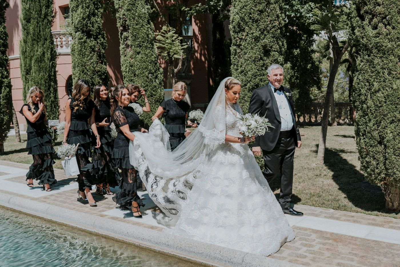 Wedding party walking along a poolside path with guests in formal attire and a flowing white bridal gown.