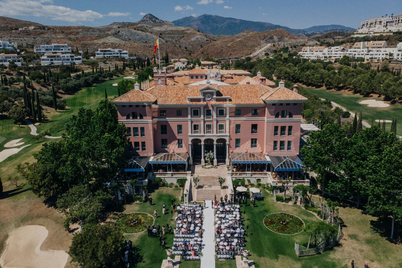 Aerial view of outdoor wedding ceremony at pink villa resort with mountain landscape and golf course in background.