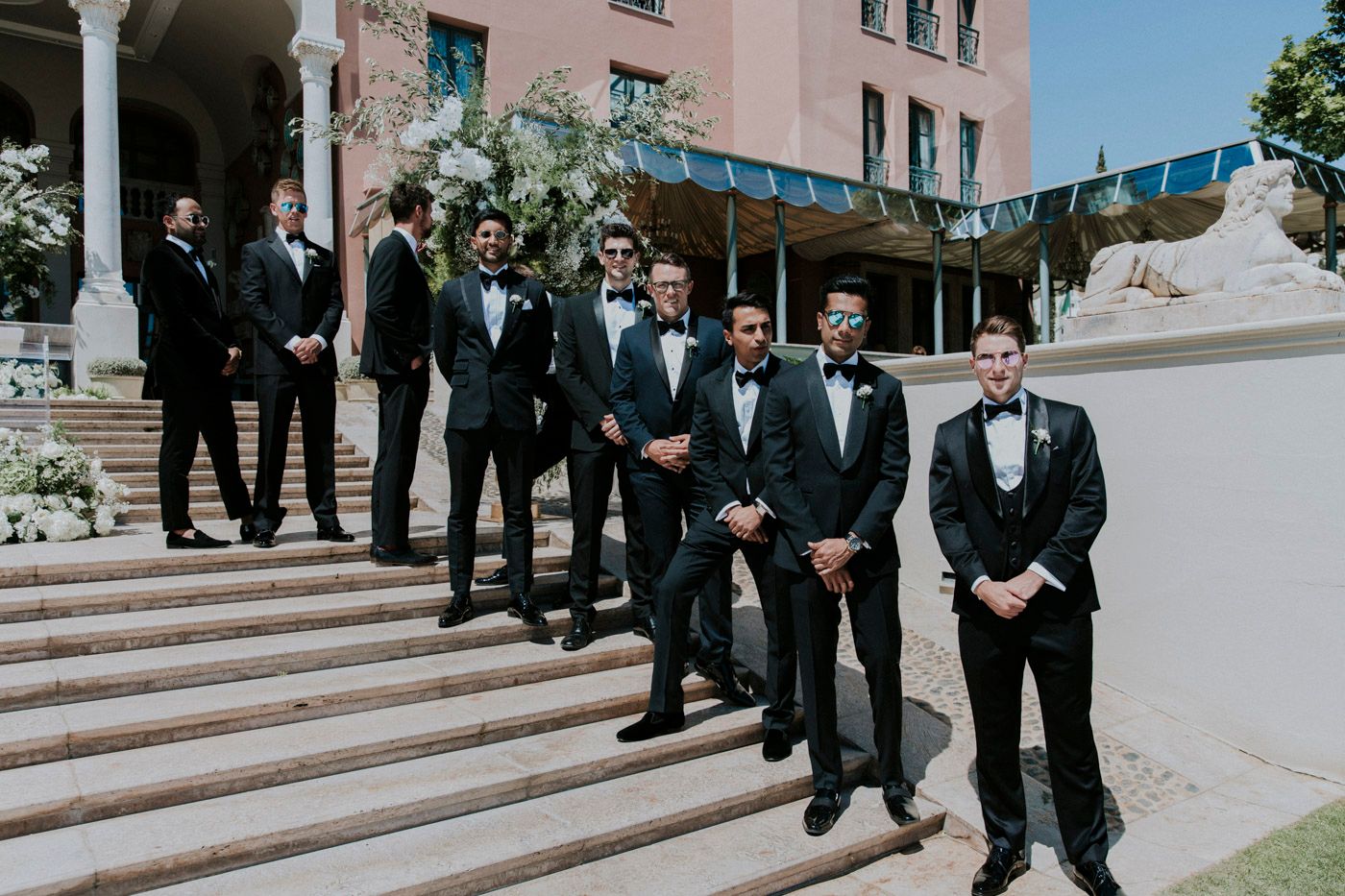 Wedding party members in formal black tuxedos lined up on stone steps of historic building.