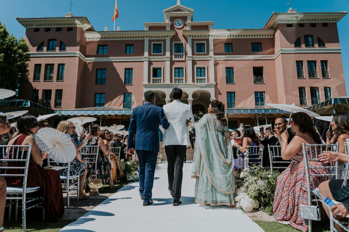 A grand outdoor wedding ceremony takes place in front of a pink historic building with guests seated on white chairs.