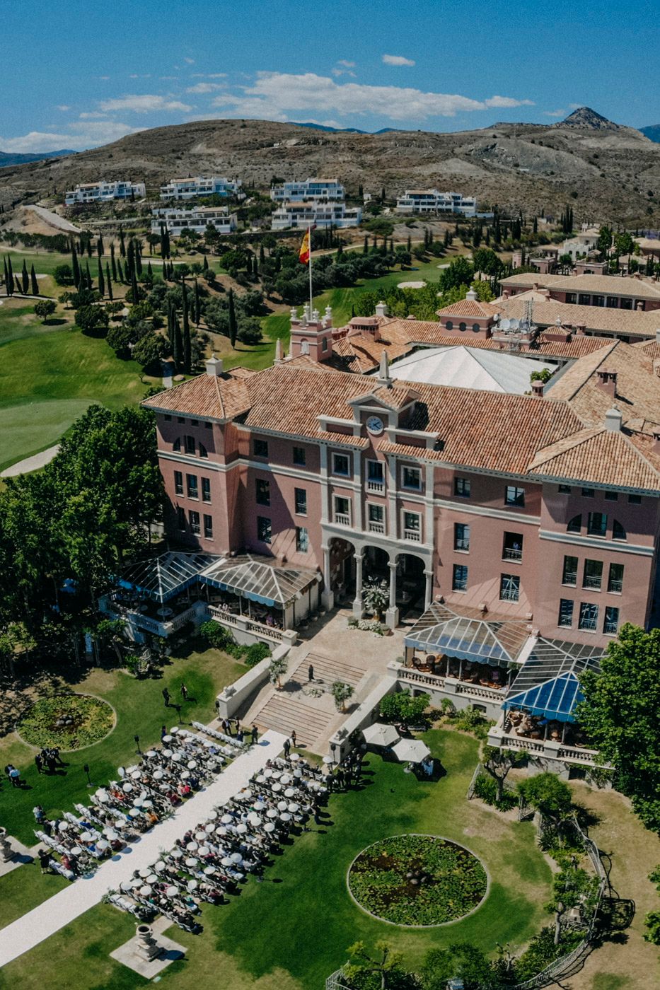 Aerial view of a pink Mediterranean-style resort building with terrace seating and mountains in the background.