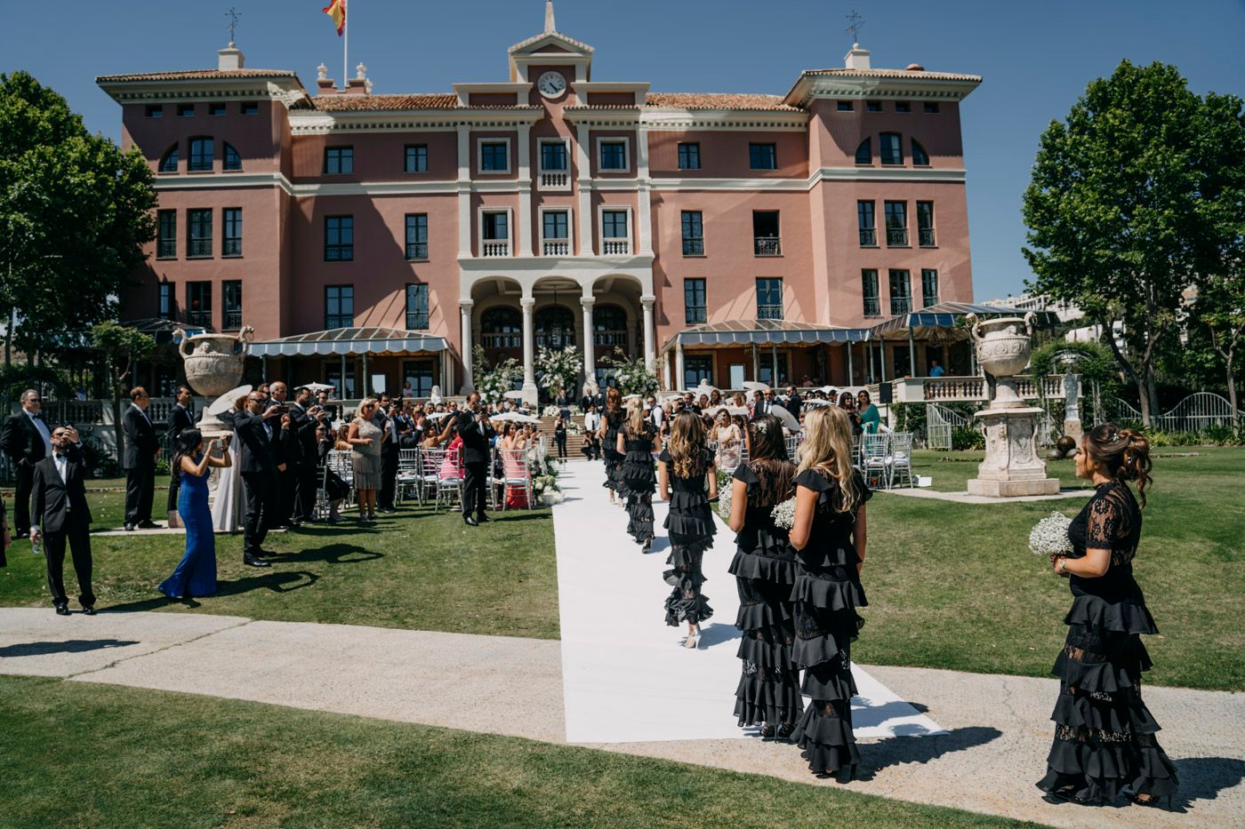 An elegant outdoor wedding procession with attendees in black gowns outside a grand pink historic hotel building.