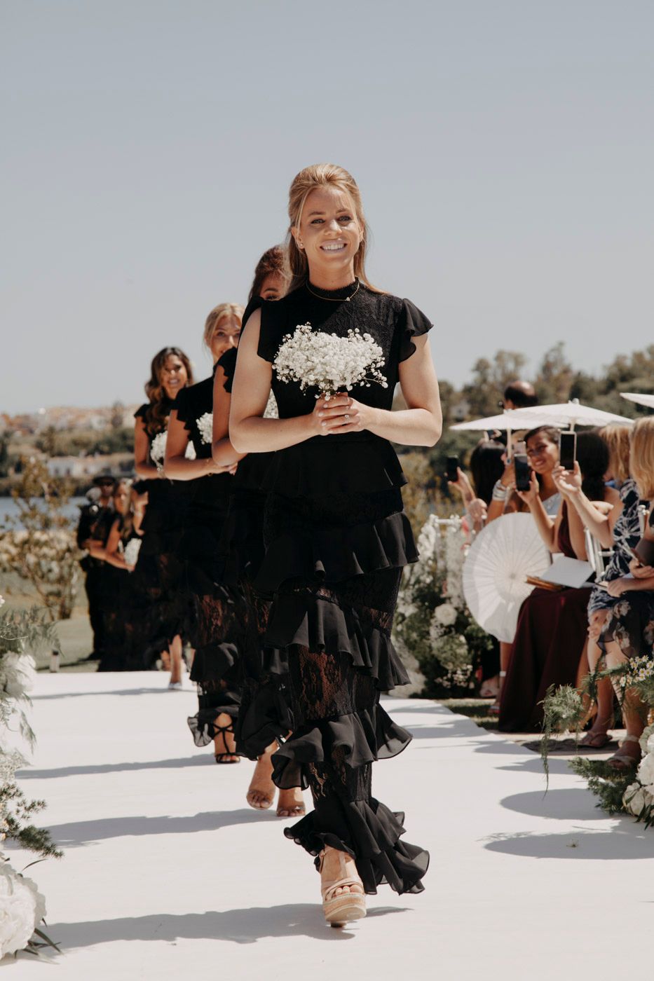 Person in a flowing black tiered dress carries a white bouquet while walking down an outdoor aisle at a wedding ceremony.