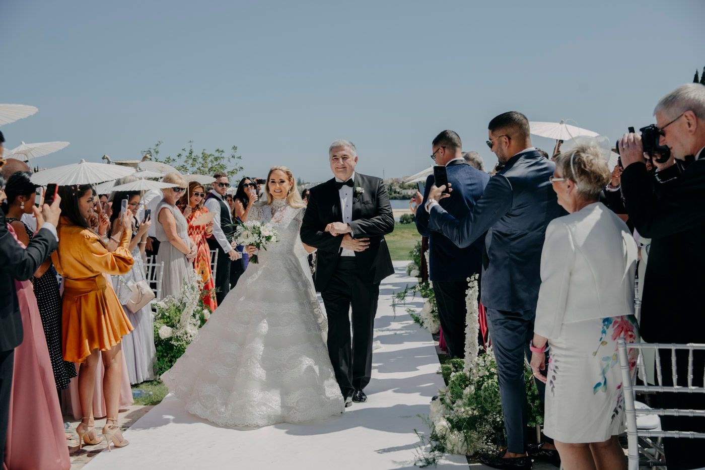 Guests line an outdoor aisle decorated with greenery as a procession makes its way through during a sunny wedding ceremony.