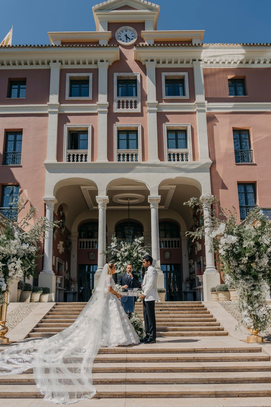 Elegant wedding ceremony on grand stone steps of pink Mediterranean-style building with white columns and clock tower.