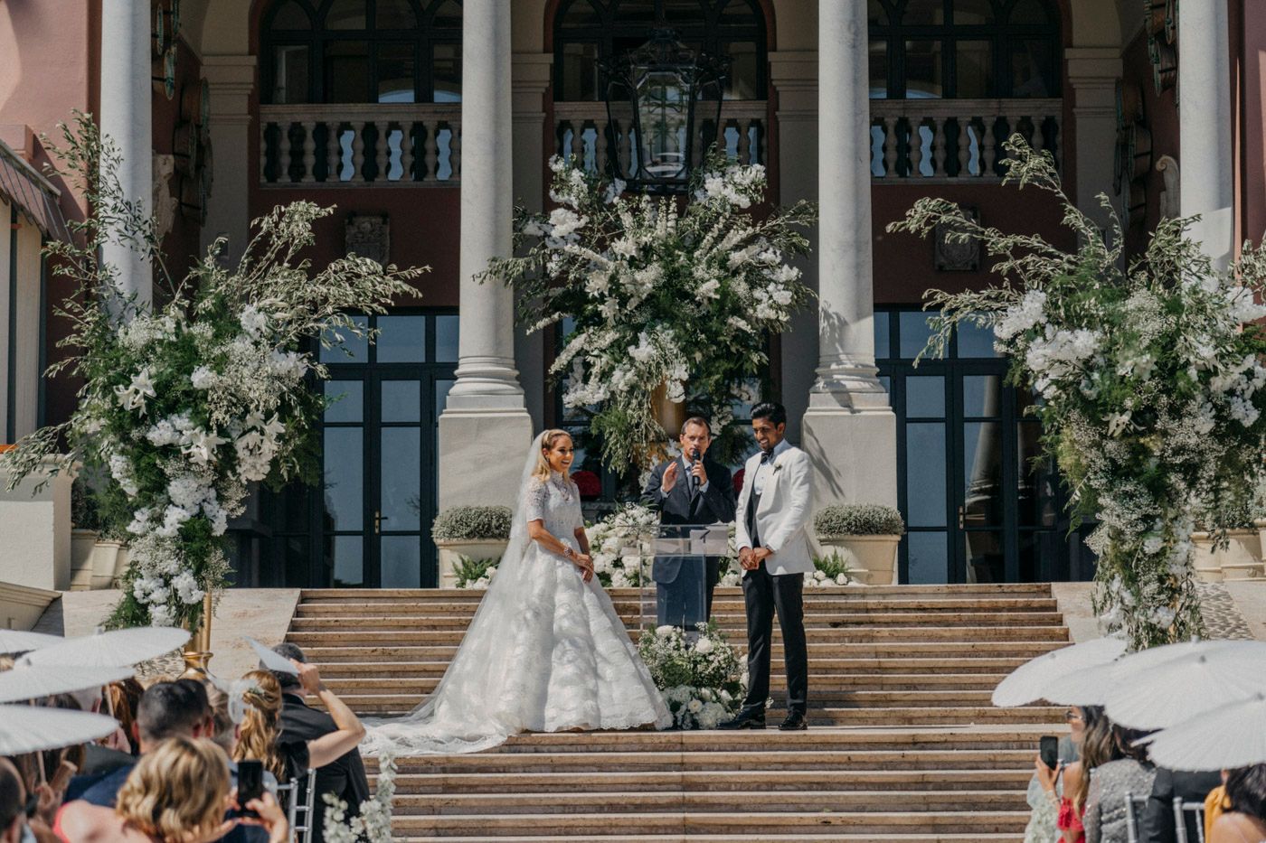 Elegant outdoor wedding ceremony on grand stone steps with white floral arrangements and classical columns.