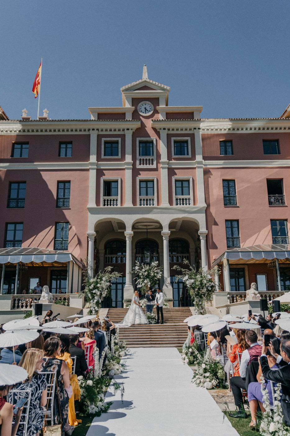 Elegant pink Mediterranean-style hotel building with grand entrance stairs decorated with floral arrangements and guests gathering.