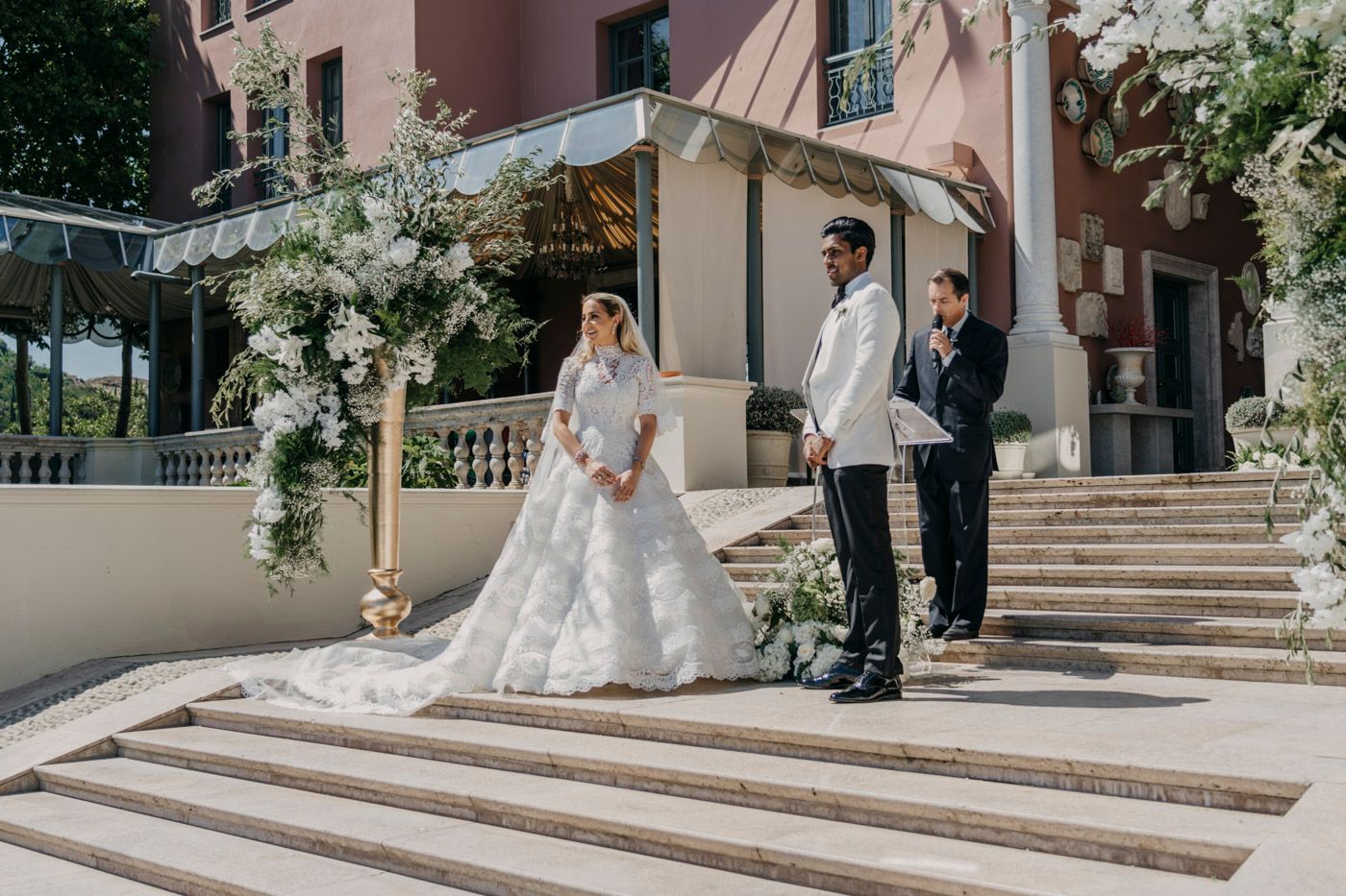 Wedding ceremony at an elegant venue with stone steps and a beautiful floral arch in natural daylight.