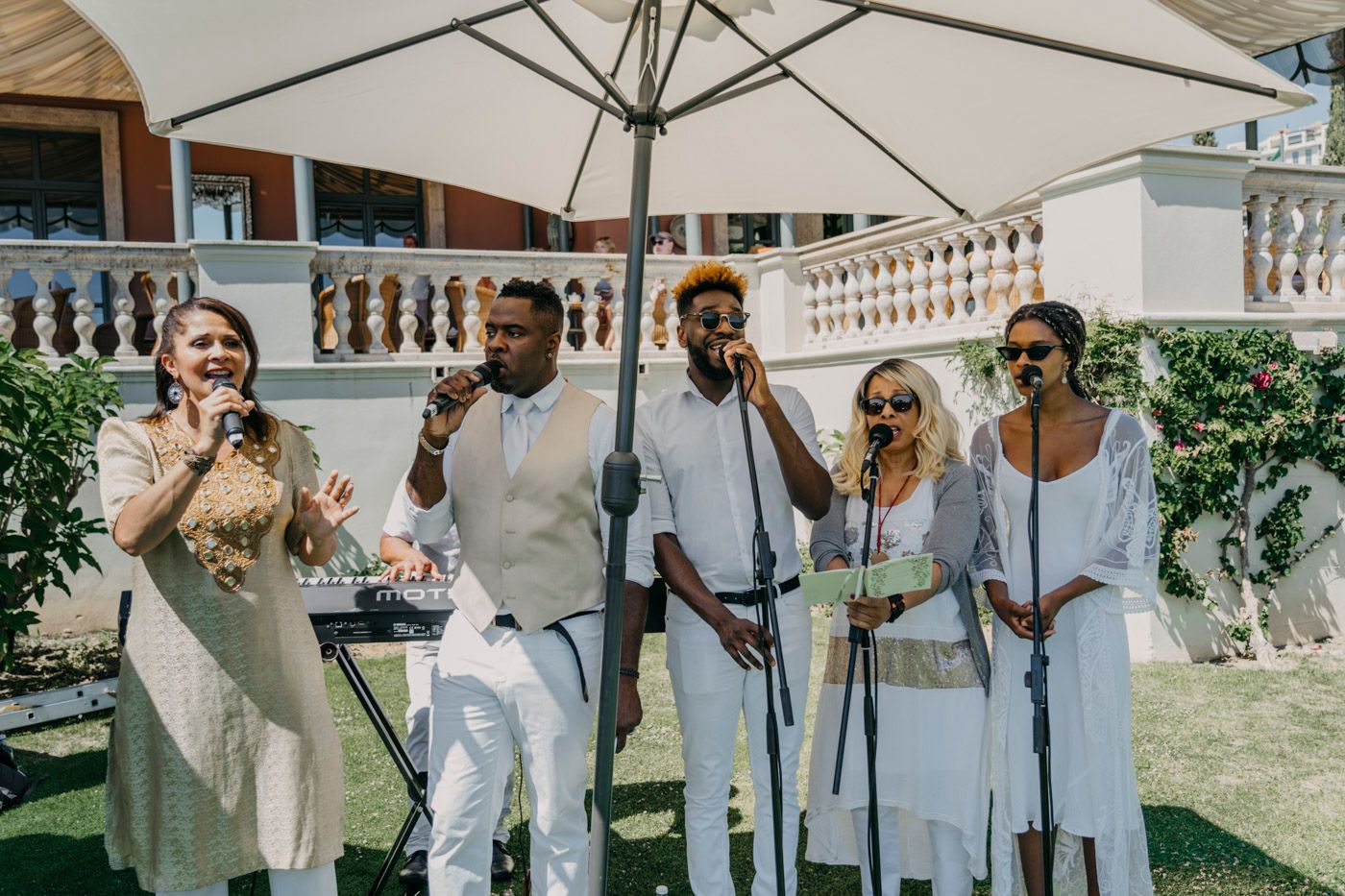 A group performs outdoors under a white umbrella wearing coordinating light colored outfits during a daytime event.
