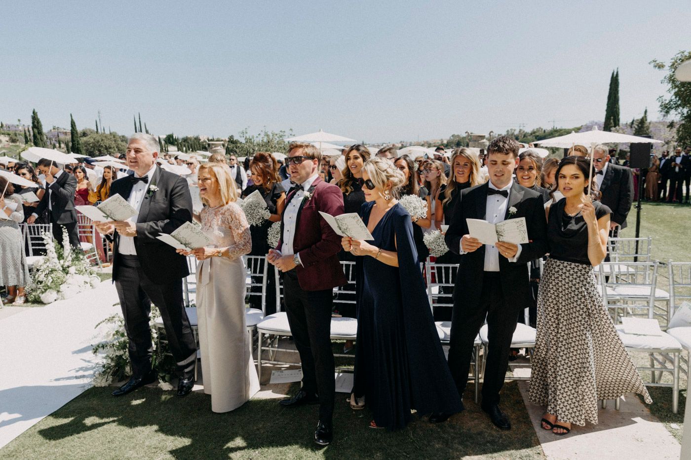 Wedding guests stand together in formal attire during an outdoor ceremony on a sunny day.