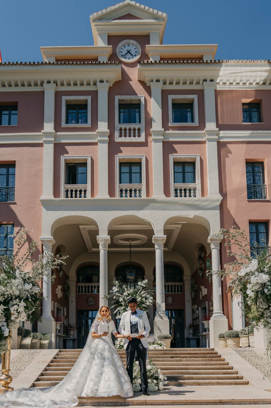 Elegant pink Mediterranean-style hotel building with classical columns and balconies decorated for a luxurious wedding ceremony.