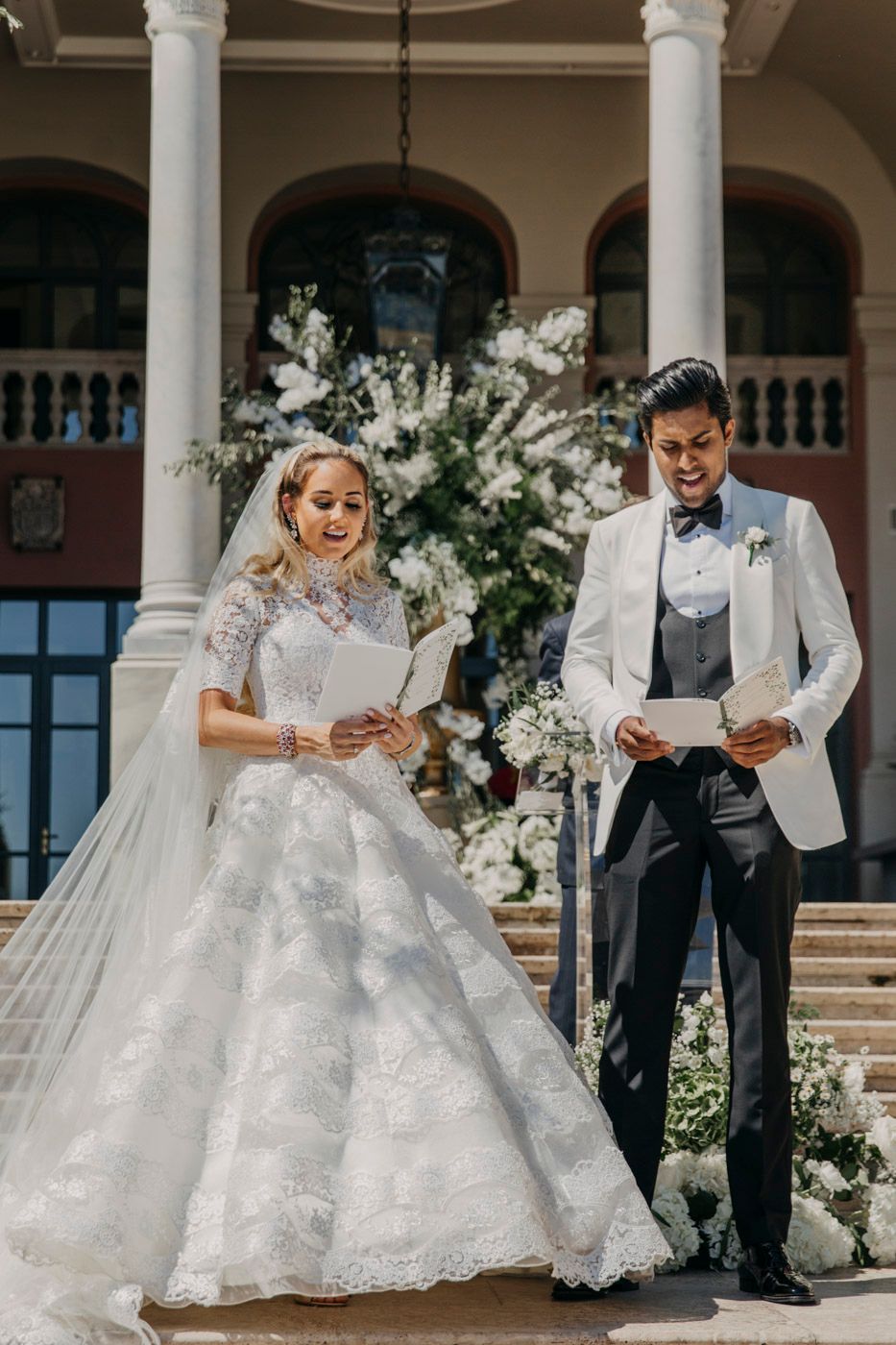Elegant wedding ceremony with couple reading vows outdoors at historic venue with white floral arrangements and classical columns.