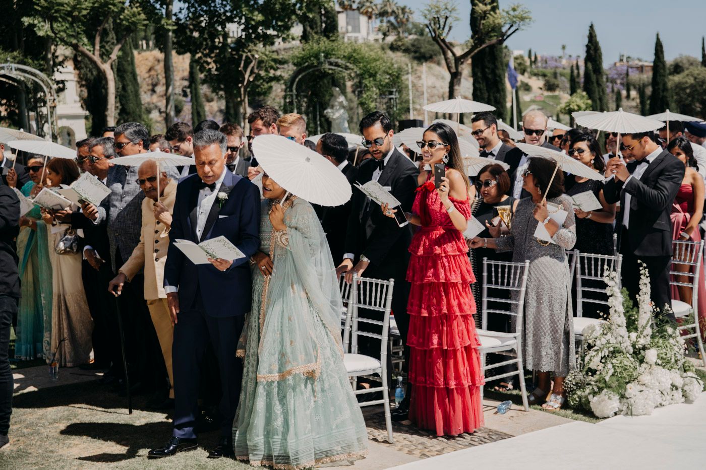 Wedding guests gather outdoors at an elegant garden ceremony, with parasols and formal attire under sunny skies.