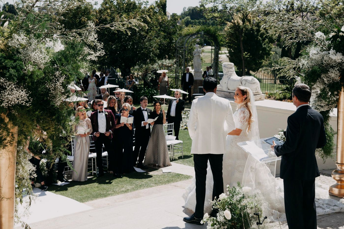 Wedding ceremony taking place in an outdoor garden setting with white floral decorations and elegant decor.