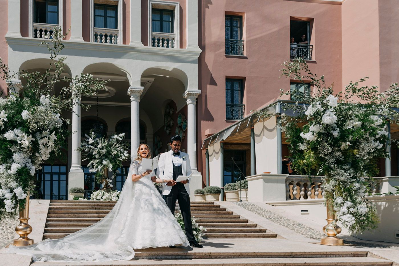 A romantic wedding moment at a pink Mediterranean-style villa with white floral arrangements and grand columns.