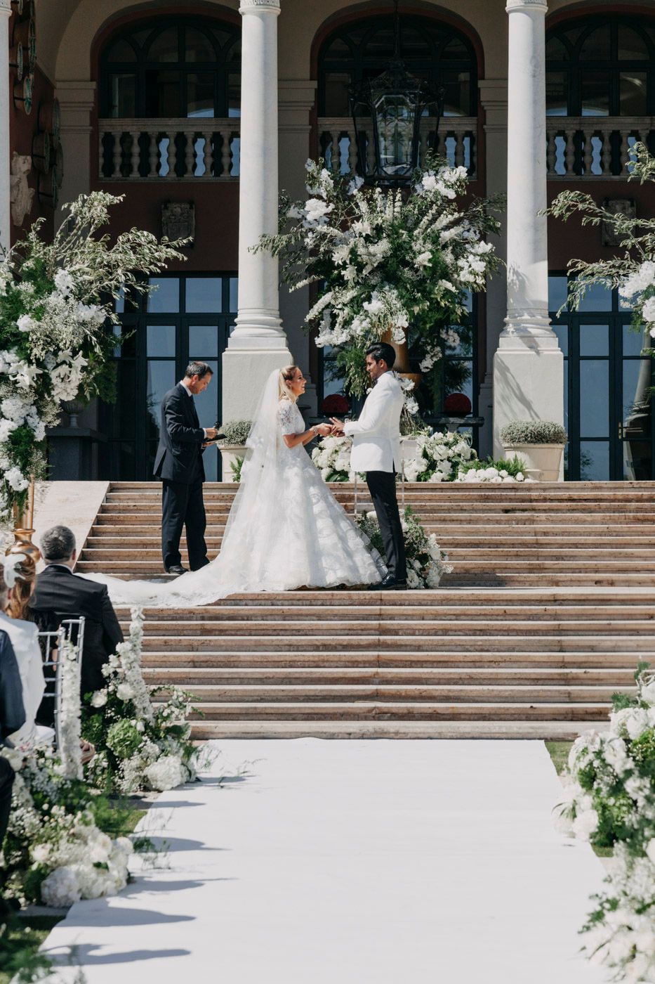 An elegant outdoor wedding ceremony on grand marble steps with white floral arrangements and columns framing the scene.