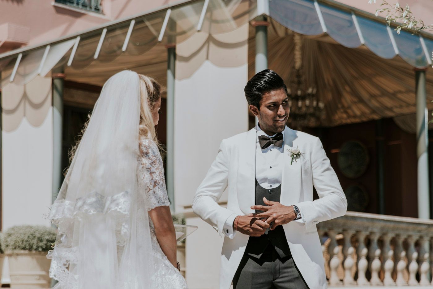 A romantic outdoor wedding scene with a couple in elegant white formalwear standing beside decorative stone columns.