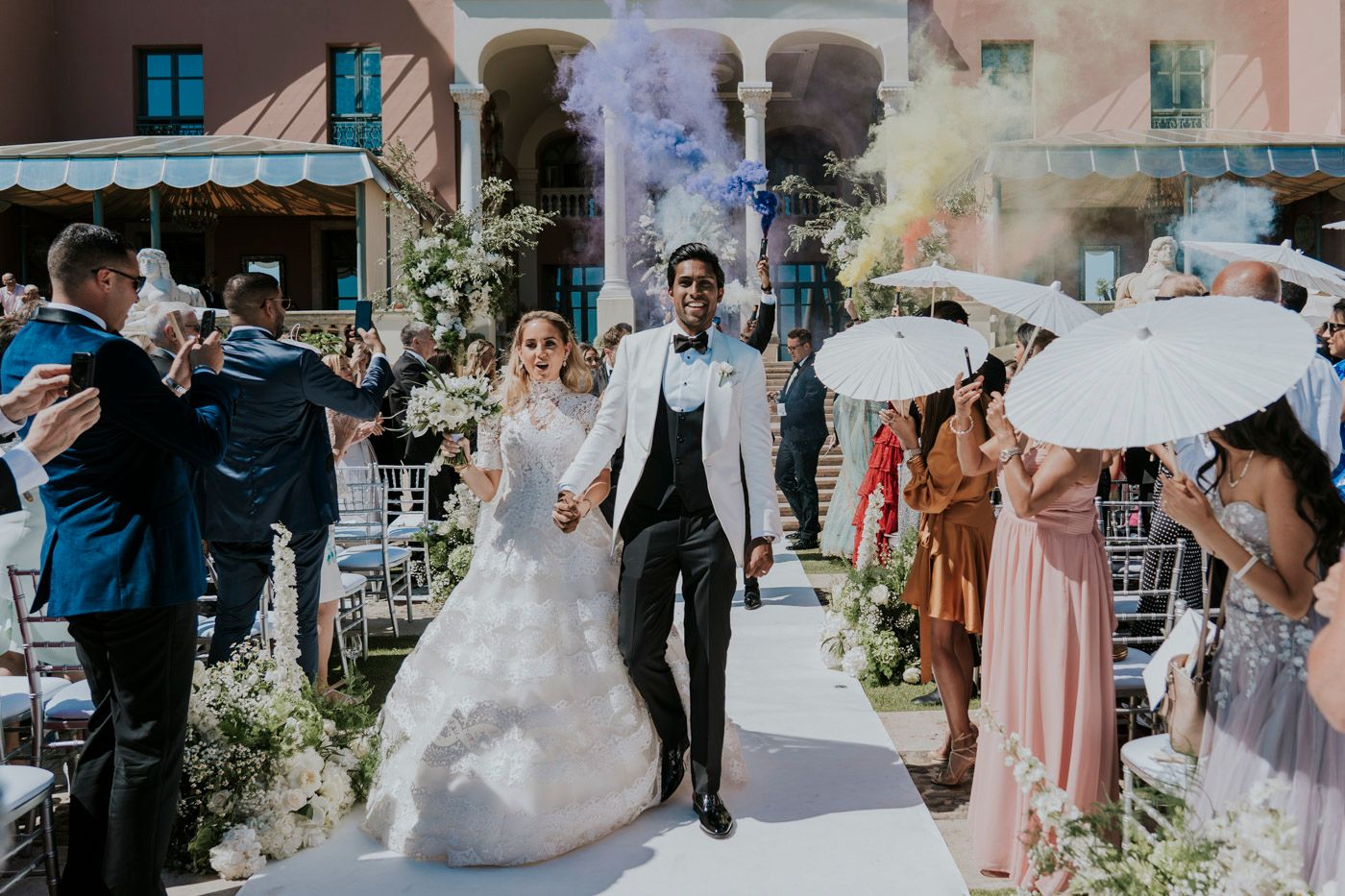 Wedding celebration with smoke effects and parasols along an outdoor aisle decorated with flowers at a villa venue.