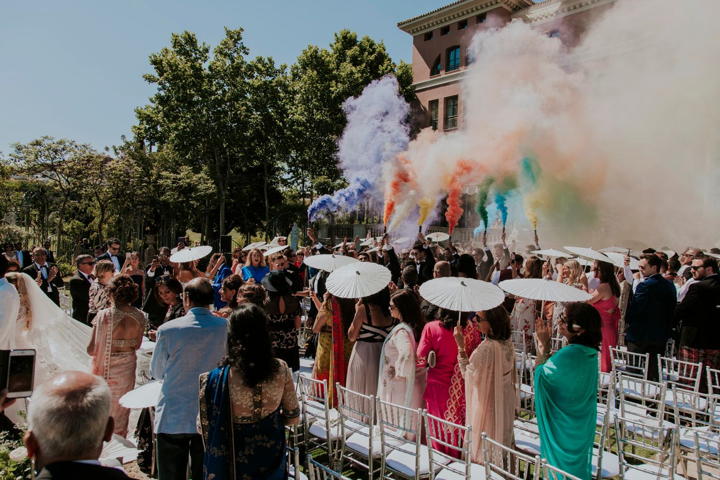 Colorful smoke bombs create vibrant clouds at an outdoor celebration with crowd and white chairs arranged for ceremony.