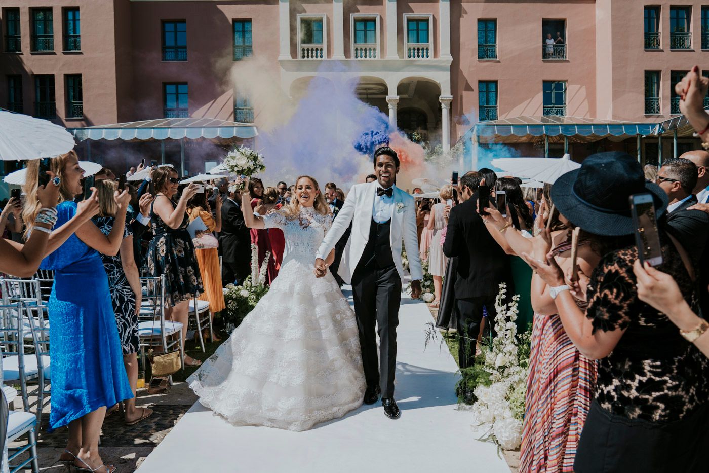 A newly wedded couple walks down an aisle filled with celebratory guests under a cloud of smoke in an outdoor ceremony.