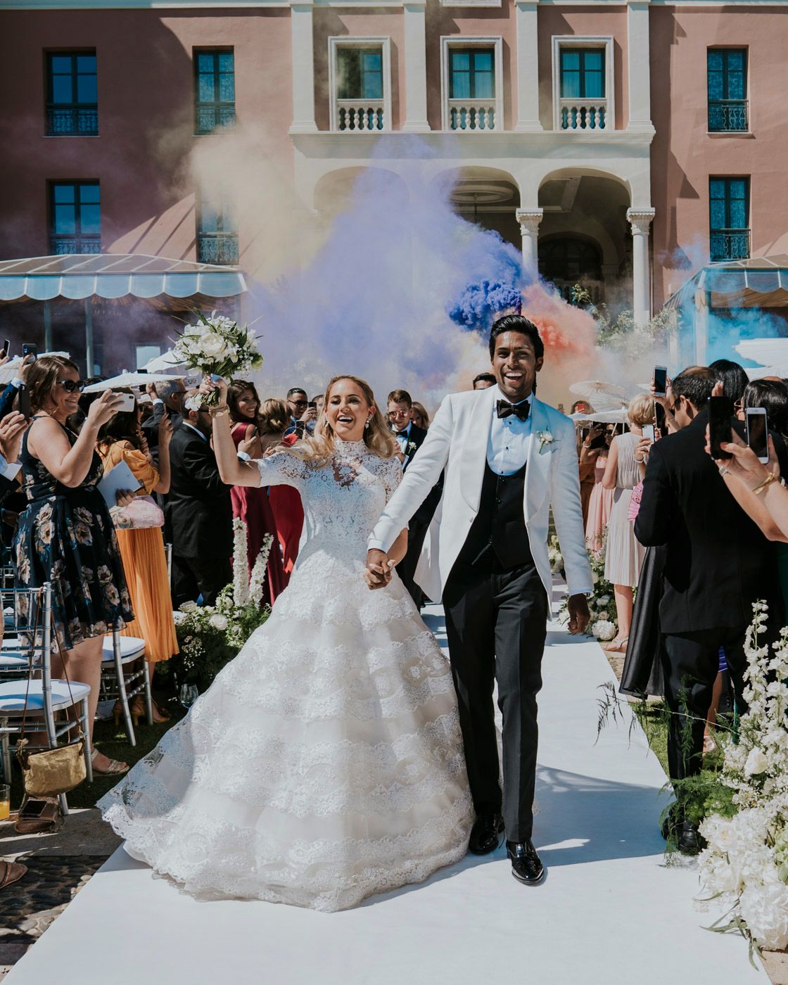 A wedding celebration with colorful smoke bombs as the couple exits along a walkway lined with floral arrangements.