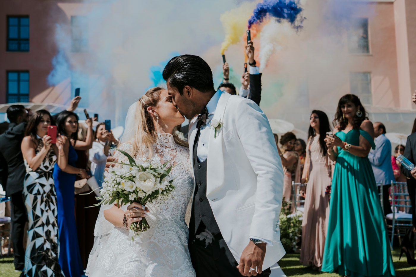 Newlyweds share a romantic kiss outdoors while guests celebrate with blue smoke flares in the background.