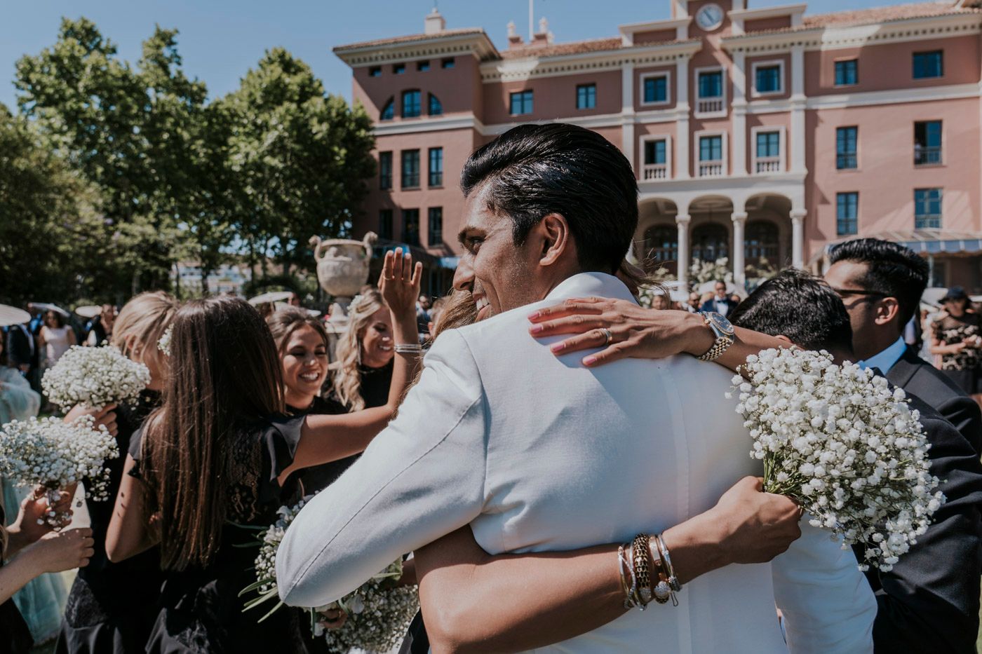 A couple embraces surrounded by white flowers and guests at an outdoor wedding celebration in front of a pink building.