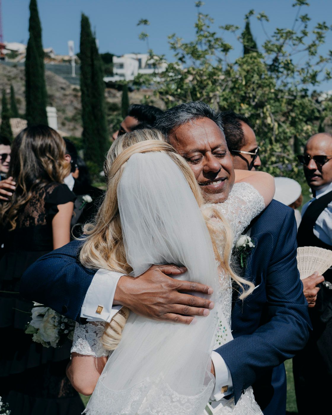 A joyful embrace between loved ones at an outdoor wedding celebration with cypress trees in the background.