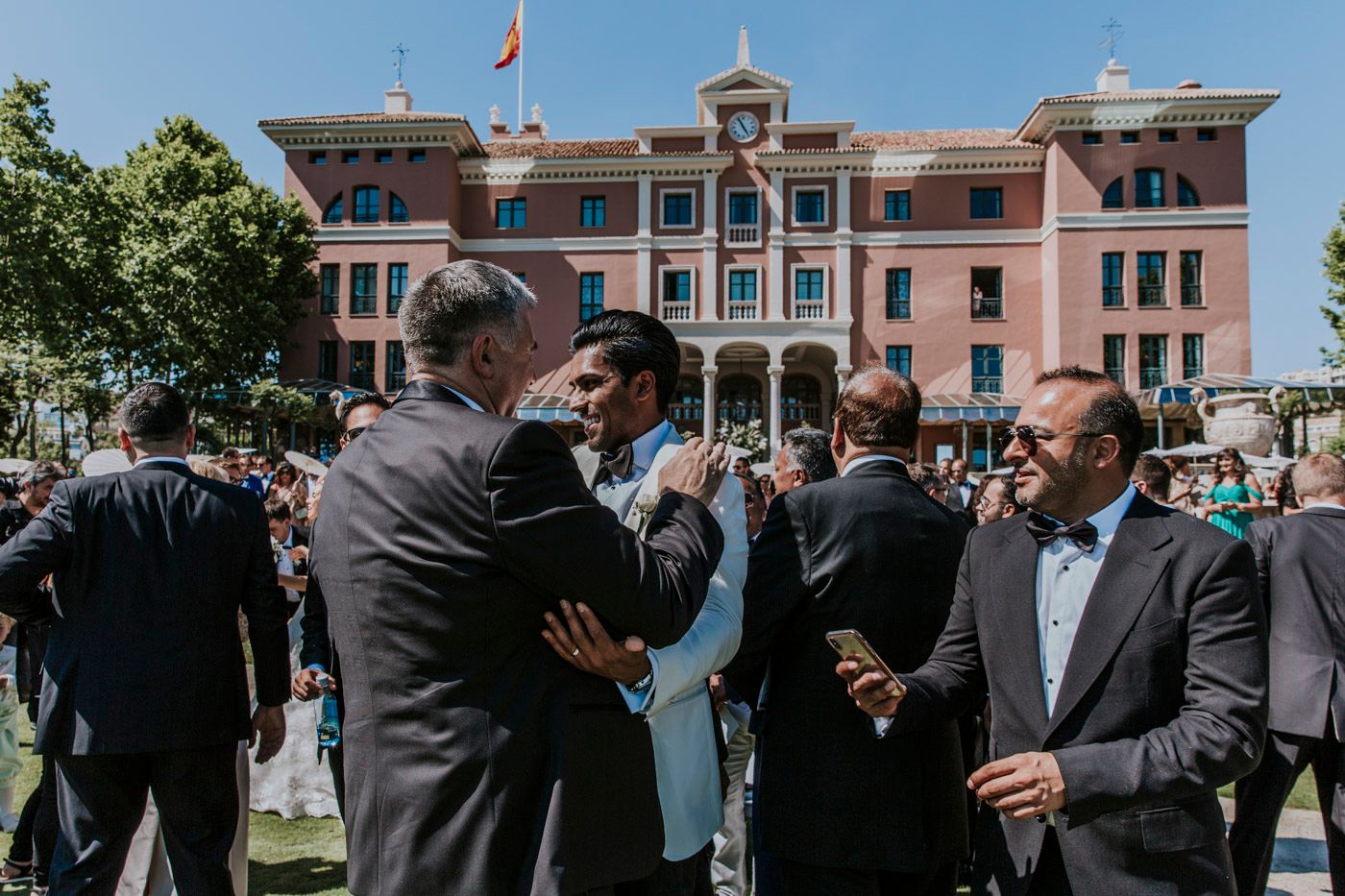 Wedding guests gather outside a pink Spanish-style building with a flag on a sunny afternoon.