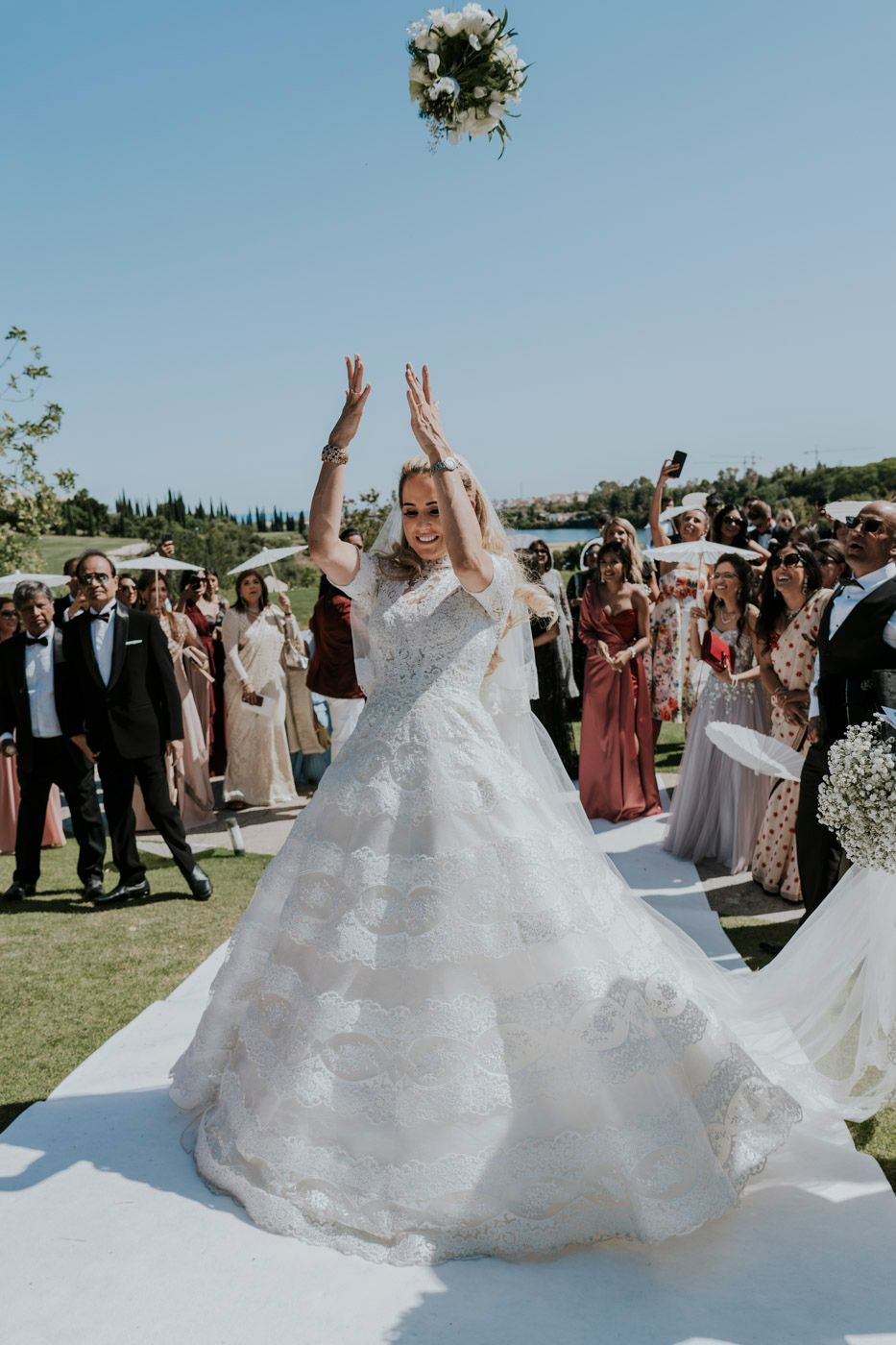 A bride in a grand white gown tosses her bouquet during an outdoor wedding ceremony.